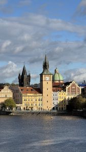 View of the Old Town Water Tower and colorful historic buildings along the Vltava River in Prague, with the Charles Bridge Tower and a green dome of St. Francis Church under a partly cloudy sky.