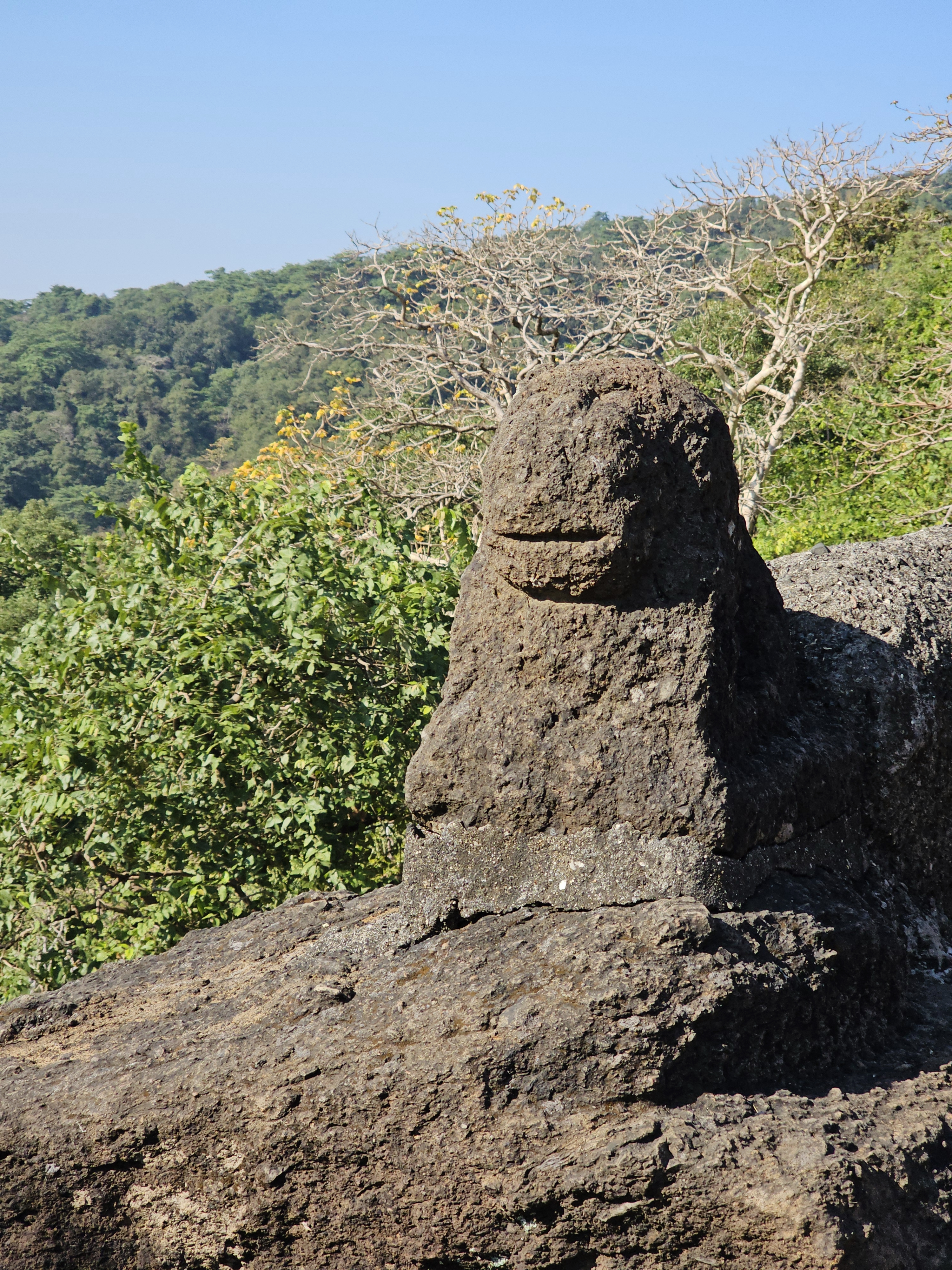 A dark, weathered stone carving resembling a smiling creature, possibly a lion, sits outdoors against a backdrop of lush green trees and a clear blue sky at Kanheri Caves, Borivali, Mumbai. 