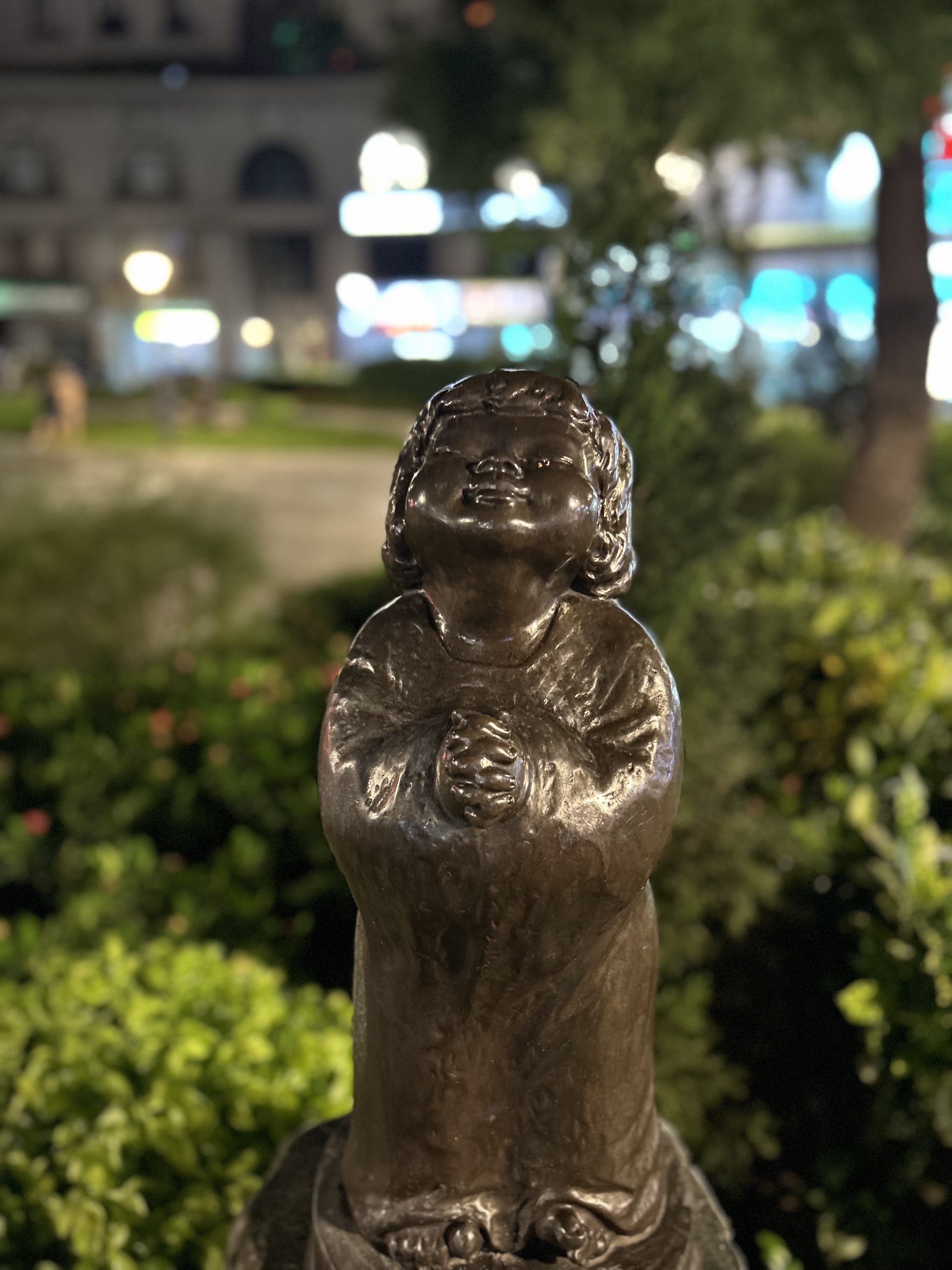 A bronze statue of a child with a round face and curly hair, depicted in a praying position with hands clasped.
