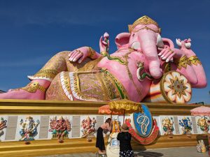 A large reclining pink statue of Lord Ganesha, adorned with gold details, set against a clear blue sky in Bangkok, Thailand.