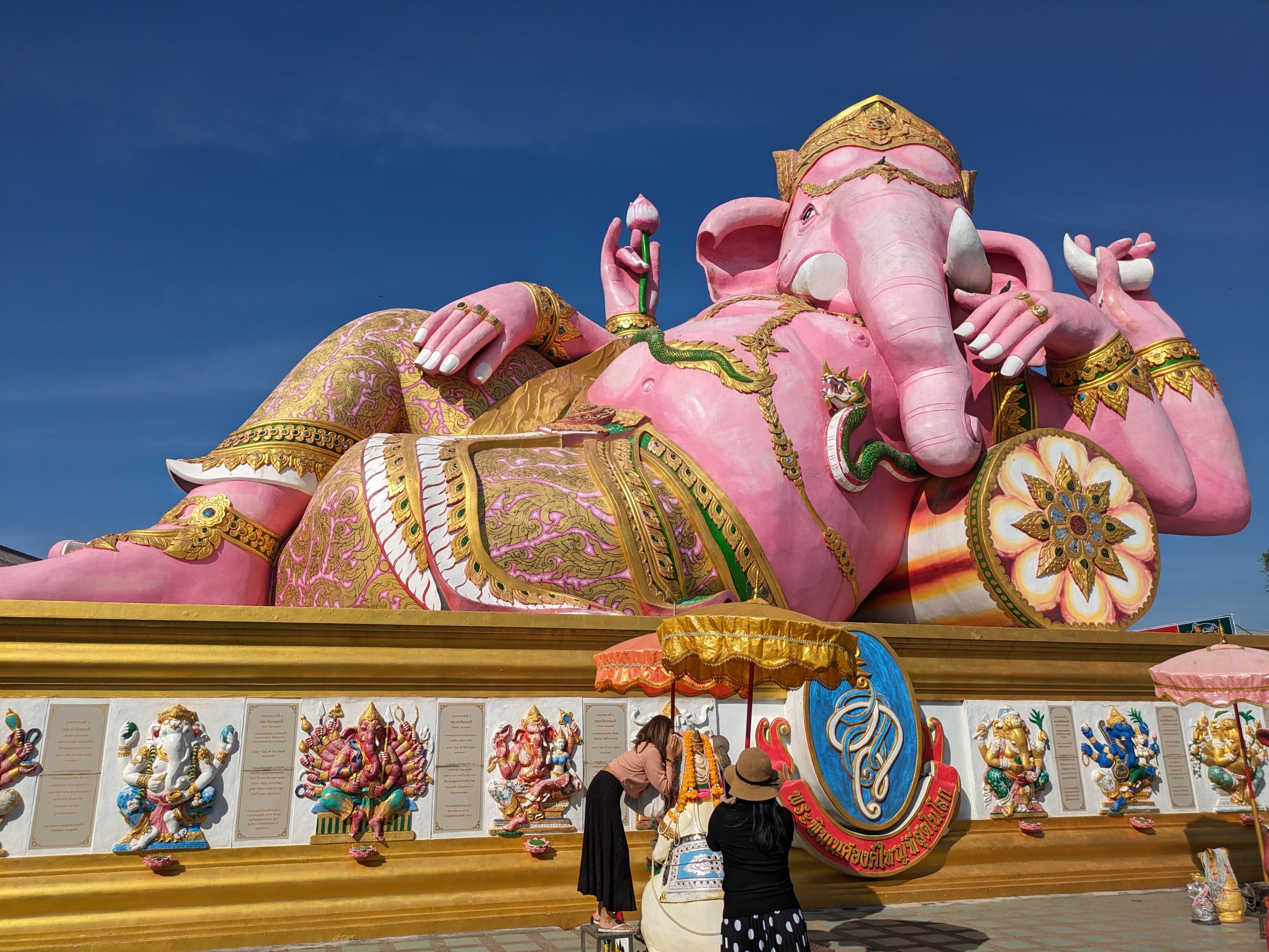 A large reclining pink statue of Lord Ganesha, adorned with gold details, set against a clear blue sky in Bangkok, Thailand.