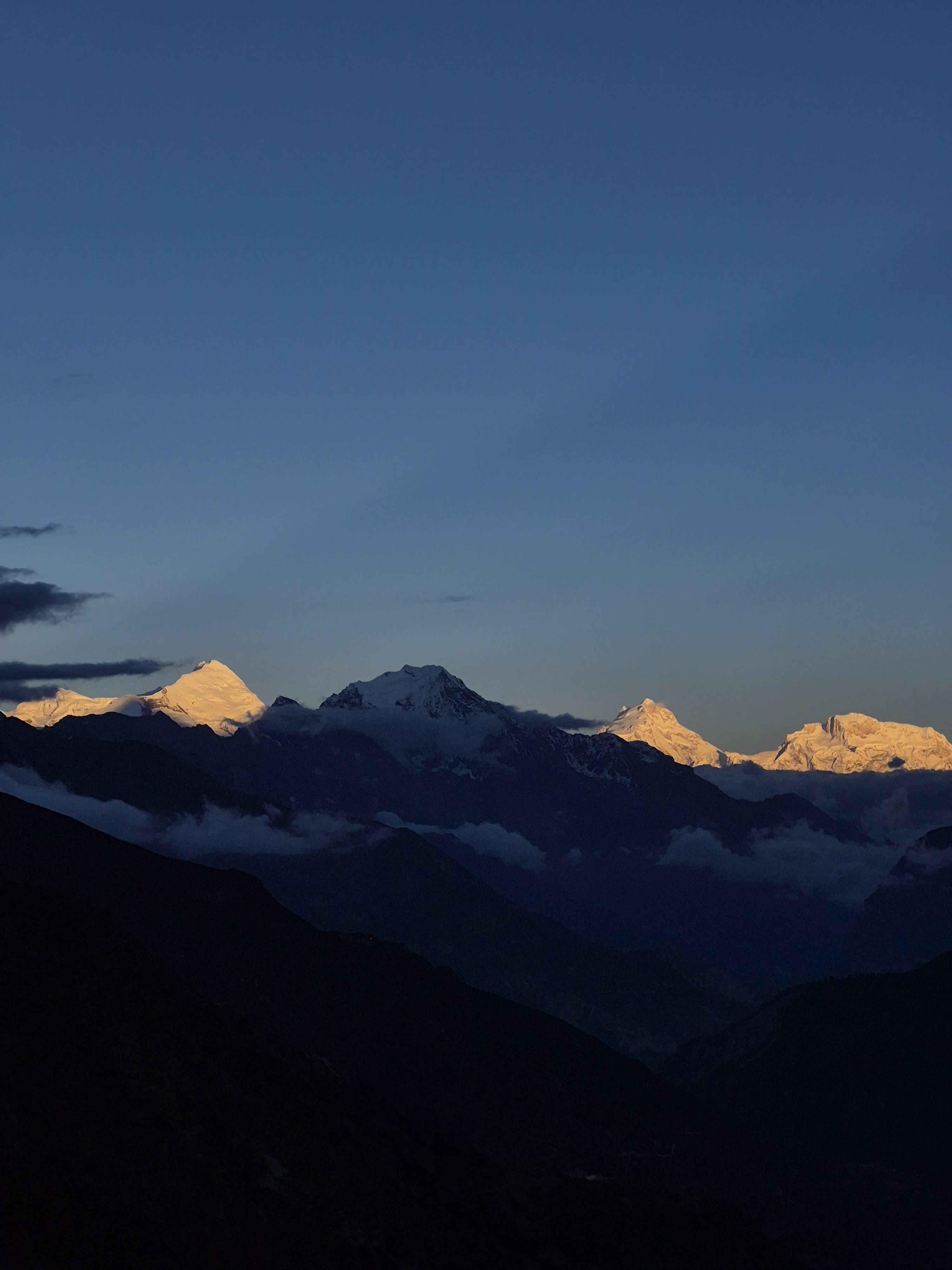 A panoramic view of snow-covered mountains under a twilight sky, with the peaks illuminated by the last rays of sunlight.