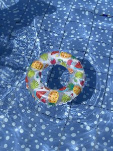 Inflatable swim ring with colorful fruit patterns floats on a blue-tiled pool. Sunlight creates rippling reflections on the water surface.