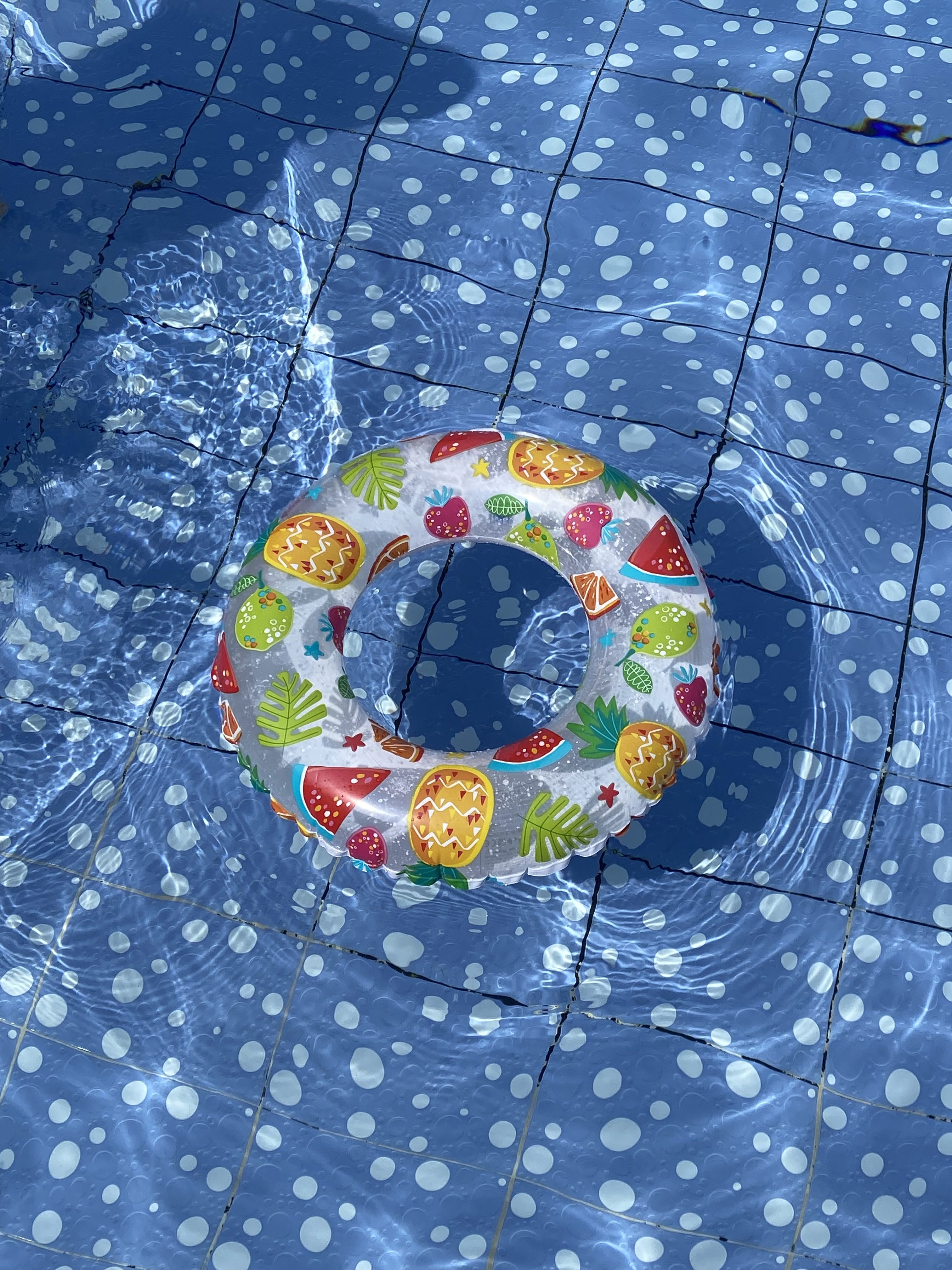 Inflatable swim ring with colorful fruit patterns floats on a blue-tiled pool. Sunlight creates rippling reflections on the water surface.