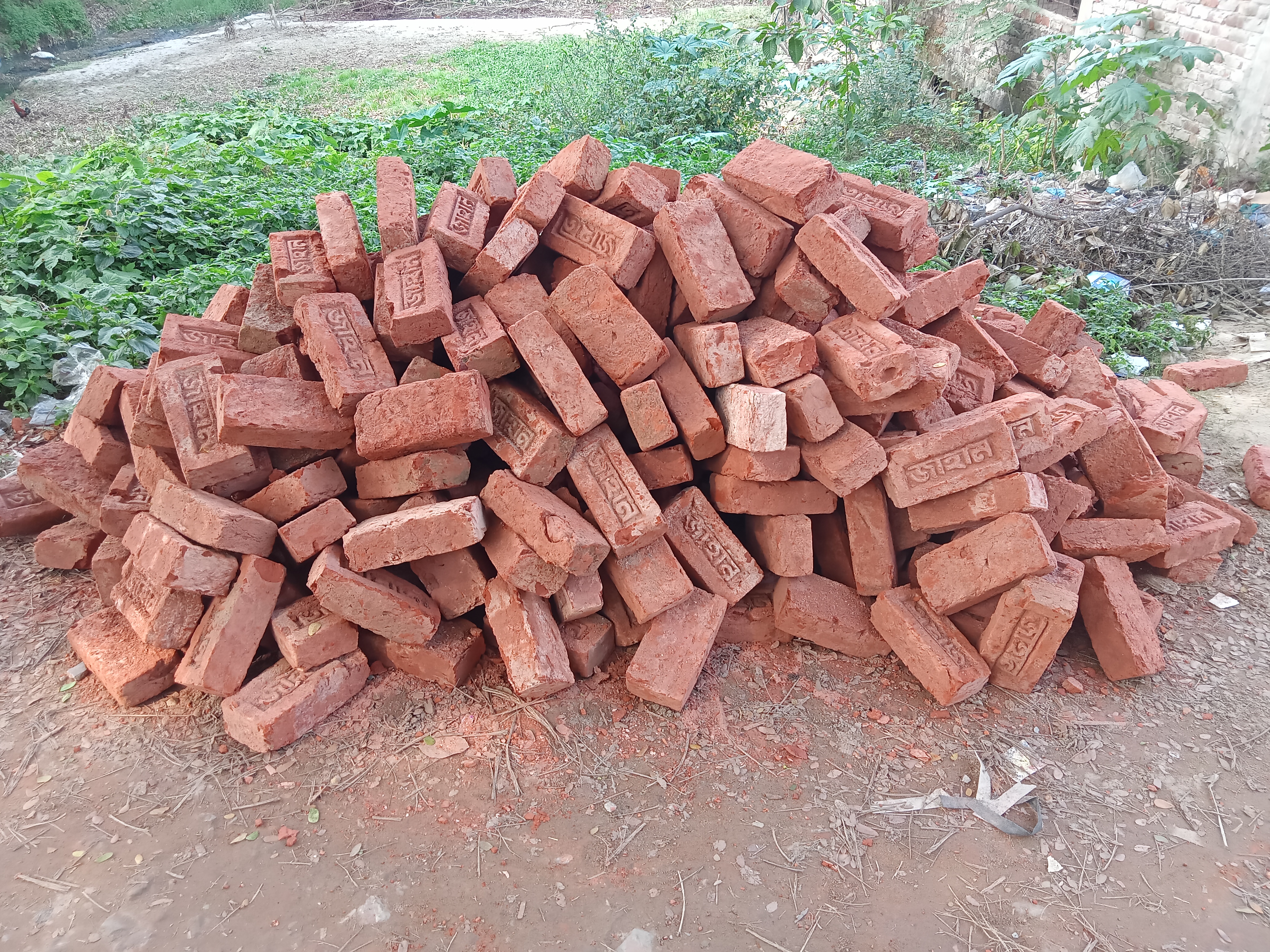 A large pile of red clay bricks stacked in a disorganized manner. The bricks have visible markings and are surrounded by green foliage and scattered debris on the ground. 