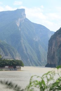 A vertical photograph capturing the Three Gorges region of the Yangtze River in China, with the river flowing between massive mountains. On the left bank, there is a small traditional building with a dark roof and white walls, surrounded by trees and sitting right at the water’s edge. The rest of the photo is dominated by tall, steep mountains.