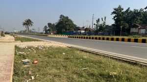 A long road with a black and yellow striped divider runs alongside a tiled sidewalk and a grassy patch where some rubbish plastics are thrown. The environment has a clear blue sky. The other side of the road has small houses and trees near the road.
