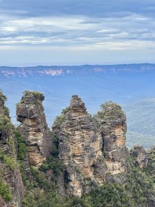 Rugged rocks surrounded by green foliage, with rolling mountains and a cloudy sky in the Blue Mountains, Australia.