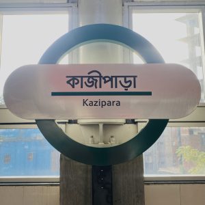 A round sign with a green and white background displays "Kazipara" in English and Bengali script. Sunlight streams through windows behind it.