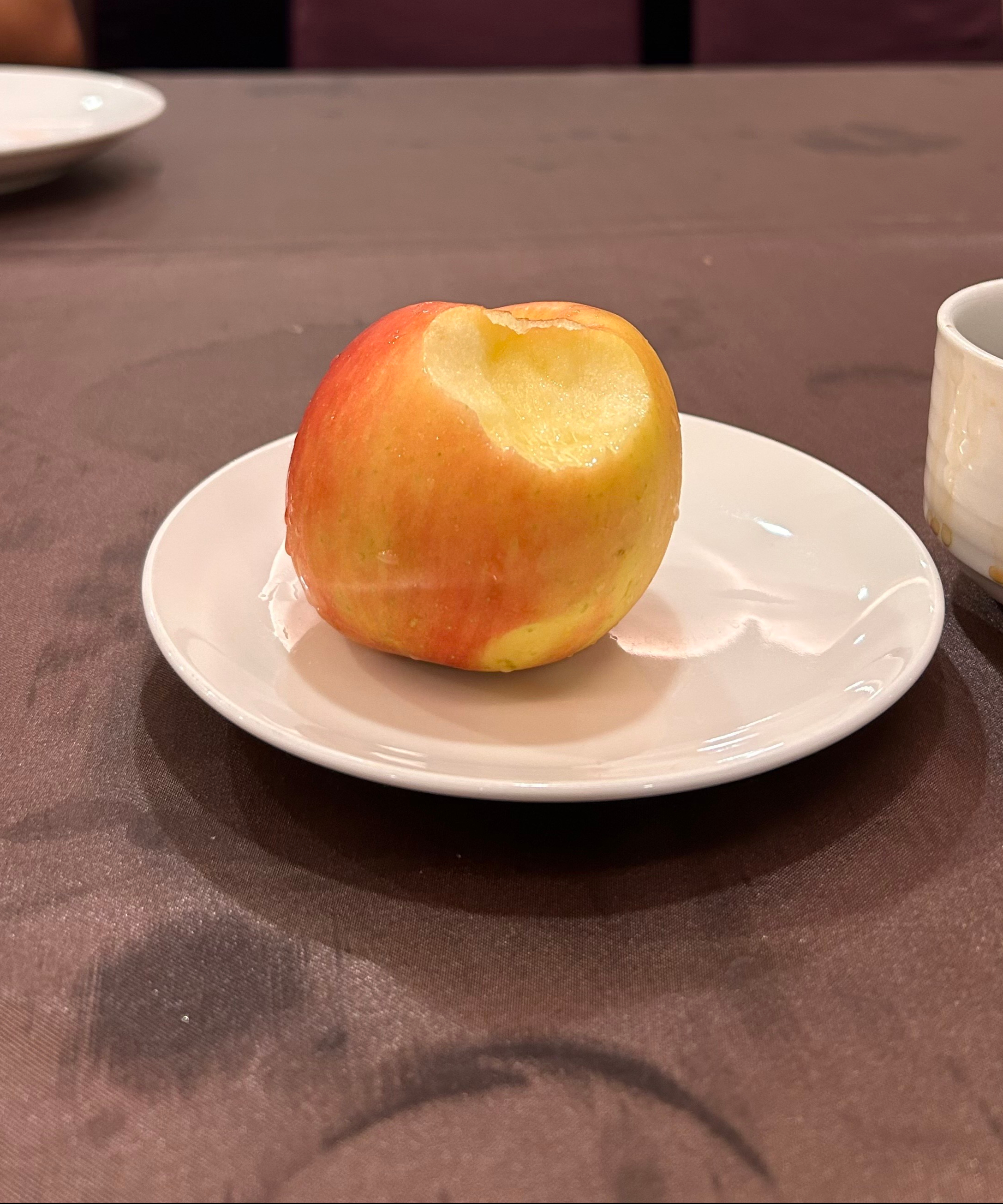 A half-eaten apple is resting on a white plate, placed on a brown tablecloth. 