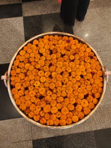 A circular metal tray is filled with vibrant orange marigold flowers, creating a dense, colorful display. 