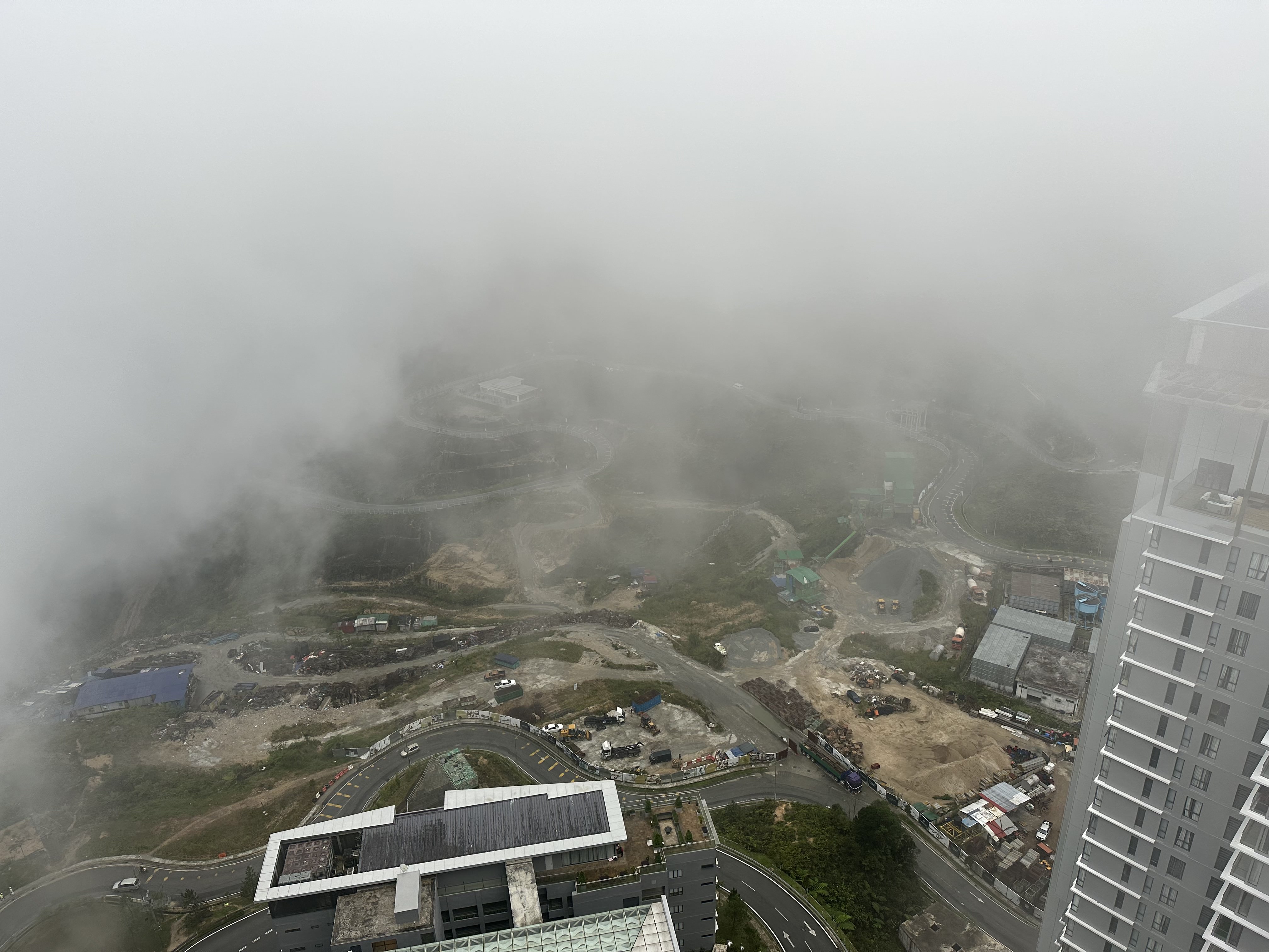 A foggy, high-altitude view of Genting Highlands, Malaysia, with clouds drifting around the buildings and winding mountain roads, revealing bits of the landscape and construction areas.