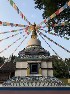 The picture shows a Buddhist stupa with a golden conical striped design at the top. Below, it is painted with the Buddha's eyes, and from the top, strings of colorful prayer flags hang down.