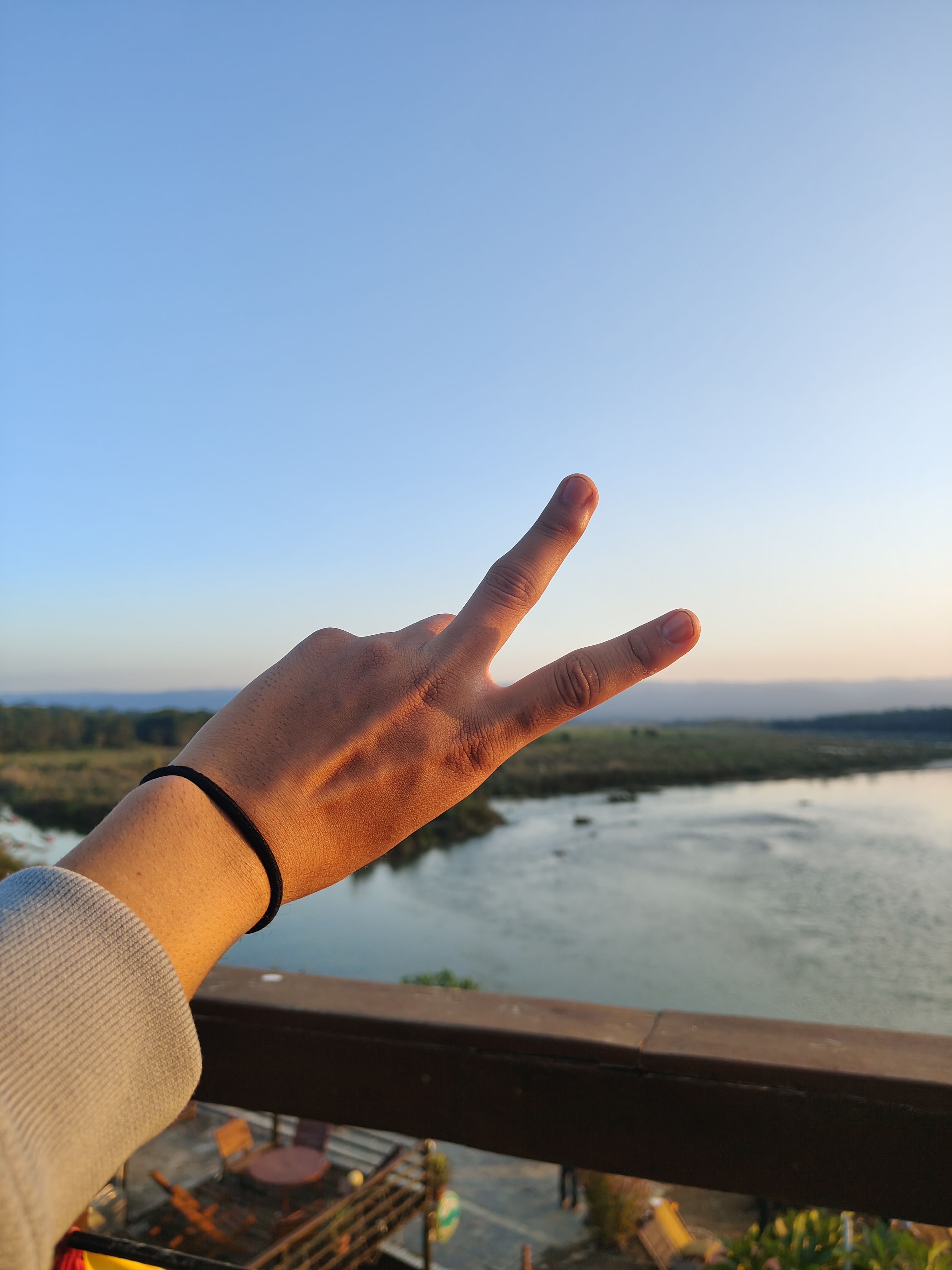 A hand making a peace sign is raised against a serene landscape featuring a river and distant hills under a clear blue sky.