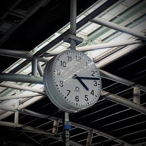 A large white clock displaying 4:13 is mounted on a metal beam structure, with a glass roof above. The setting suggests a modern train station.