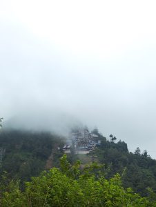 A misty mountain landscape featuring a partially obscured building complex situated on a slope.