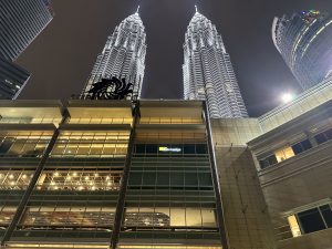 This image shows the Petronas Twin Towers in Kuala Lumpur at night, brightly illuminated against the dark sky, with the Suria KLCC mall and a Kinokuniya bookstore visible in the foreground.