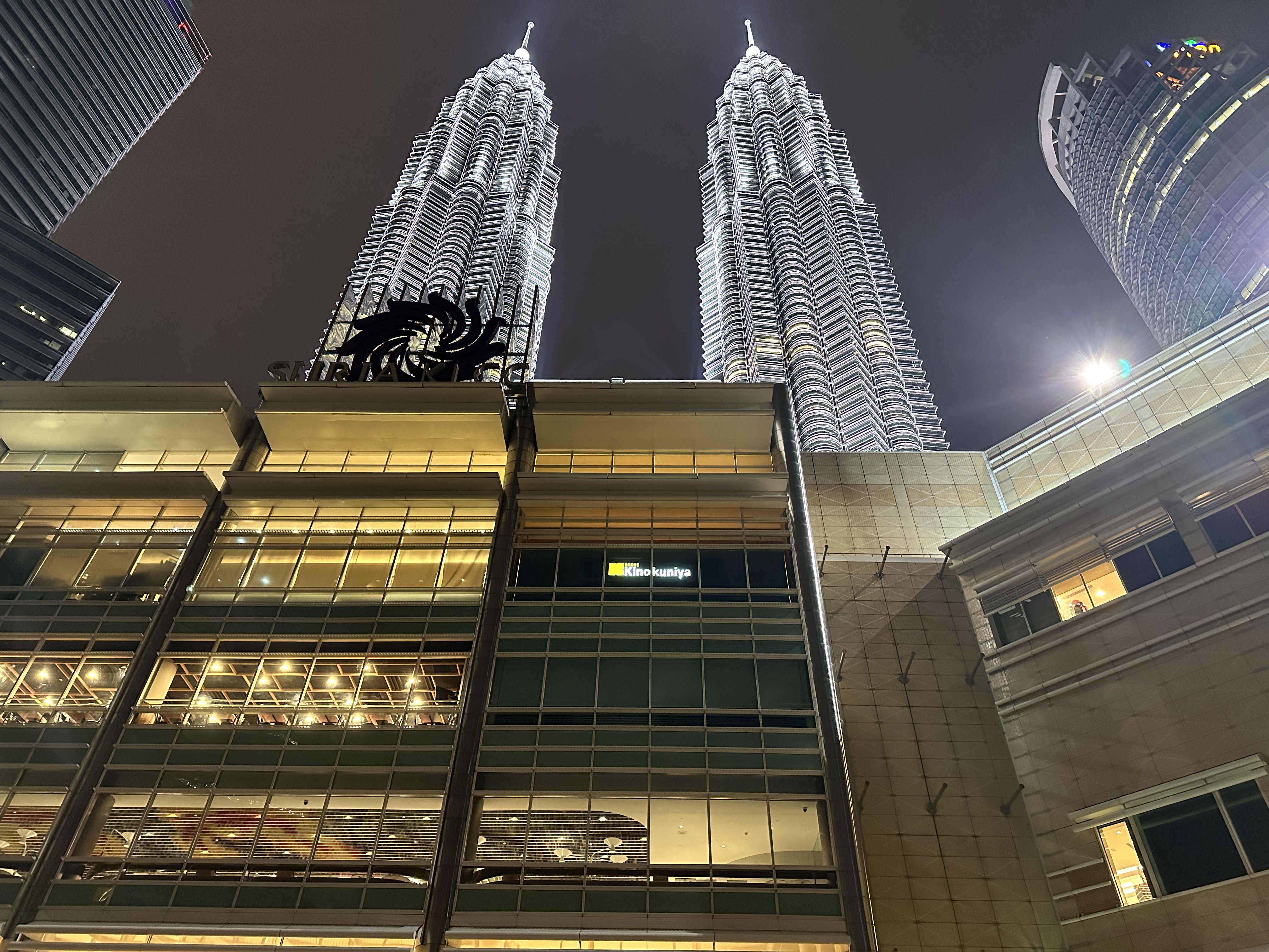 This image shows the Petronas Twin Towers in Kuala Lumpur at night, brightly illuminated against the dark sky, with the Suria KLCC mall and a Kinokuniya bookstore visible in the foreground.