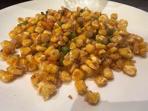 A white plate is piled high with seasoned, pan-fried sweet corn kernels mixed with chopped onions, green bell peppers, and carrots.