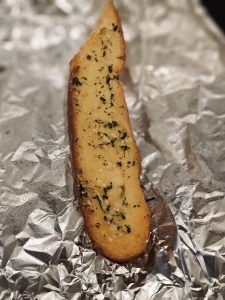 A single slice of garlic bread resting on silver foil, taken near a cafe in Asalpha, Mumbai. The toasted edges, herbs, and buttery texture make it look simple and delicious.