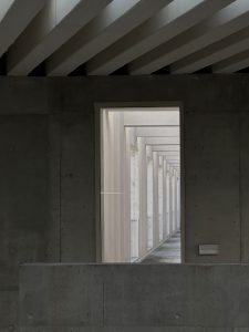 A rectangular window framed by concrete walls, showcasing a corridor lined with tall, slender columns and soft, natural lighting. The perspective creates a sense of depth as the corridor extends into the distance, with minimalistic design elements and a smooth floor visible.