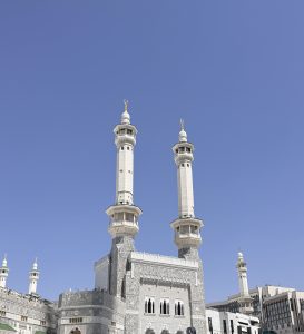Two white minarets of the Grand Mosque in Mecca rise against a blue sky, with pilgrims in white below.
