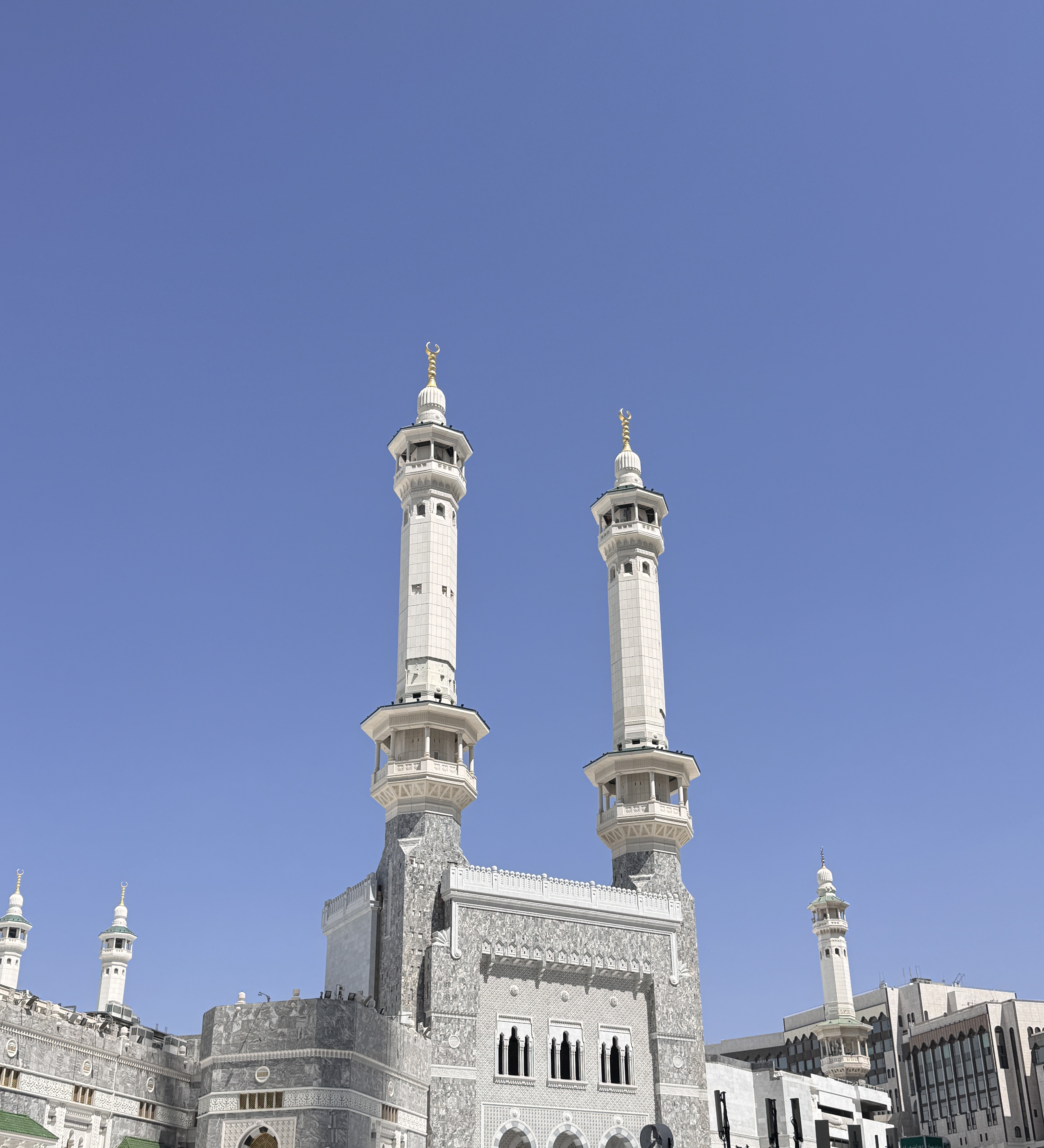 Two white minarets of the Grand Mosque in Mecca rise against a blue sky, with pilgrims in white below.