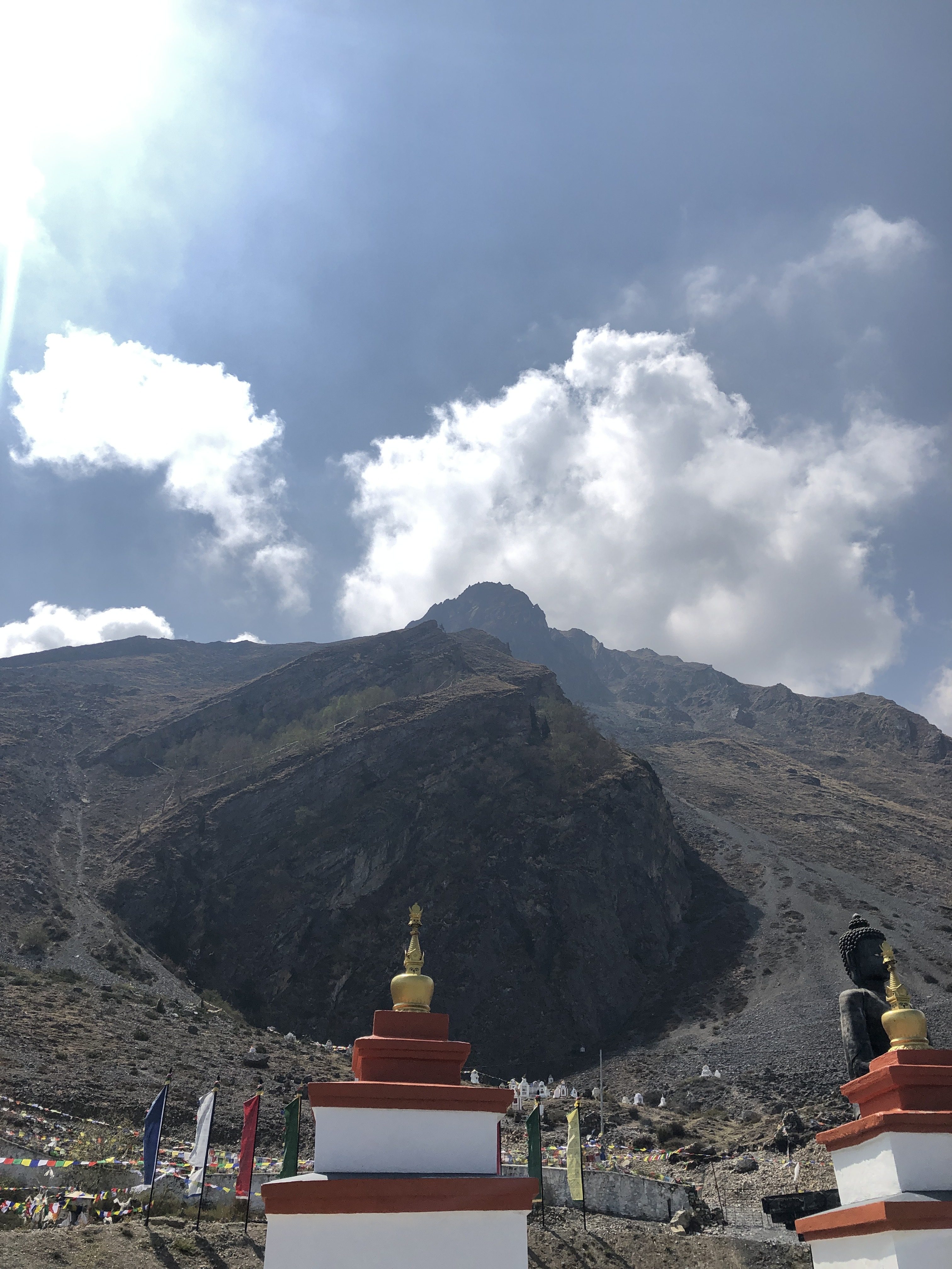 A scenic view featuring two white stupas with golden spires in the foreground, framed by a rocky mountainside that rises sharply behind them.
