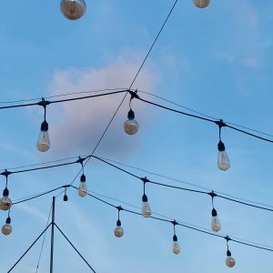 String lights hang against a clear blue sky with wispy clouds, creating a serene, outdoor ambiance. The bulbs are off, adding a rustic charm.