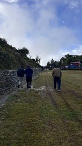 Three individuals are walking along a grassy path bordered by a stone wall, with lush greenery and low trees on one side. 