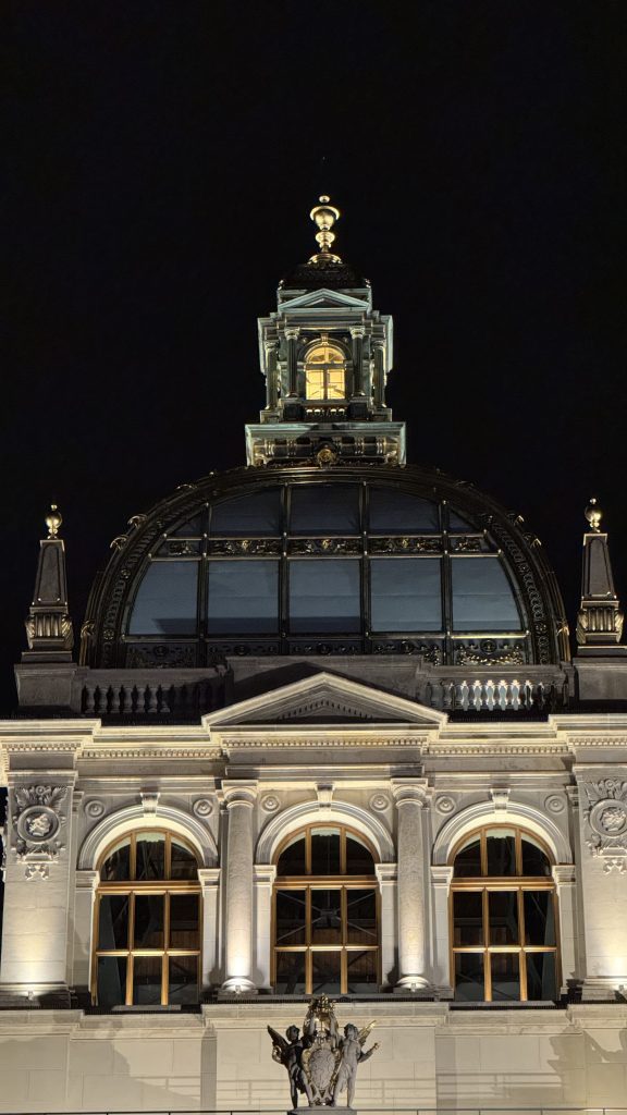 Prague historic building at night with an illuminated dome, arched windows, classical columns, and a glowing lantern in the cupola.