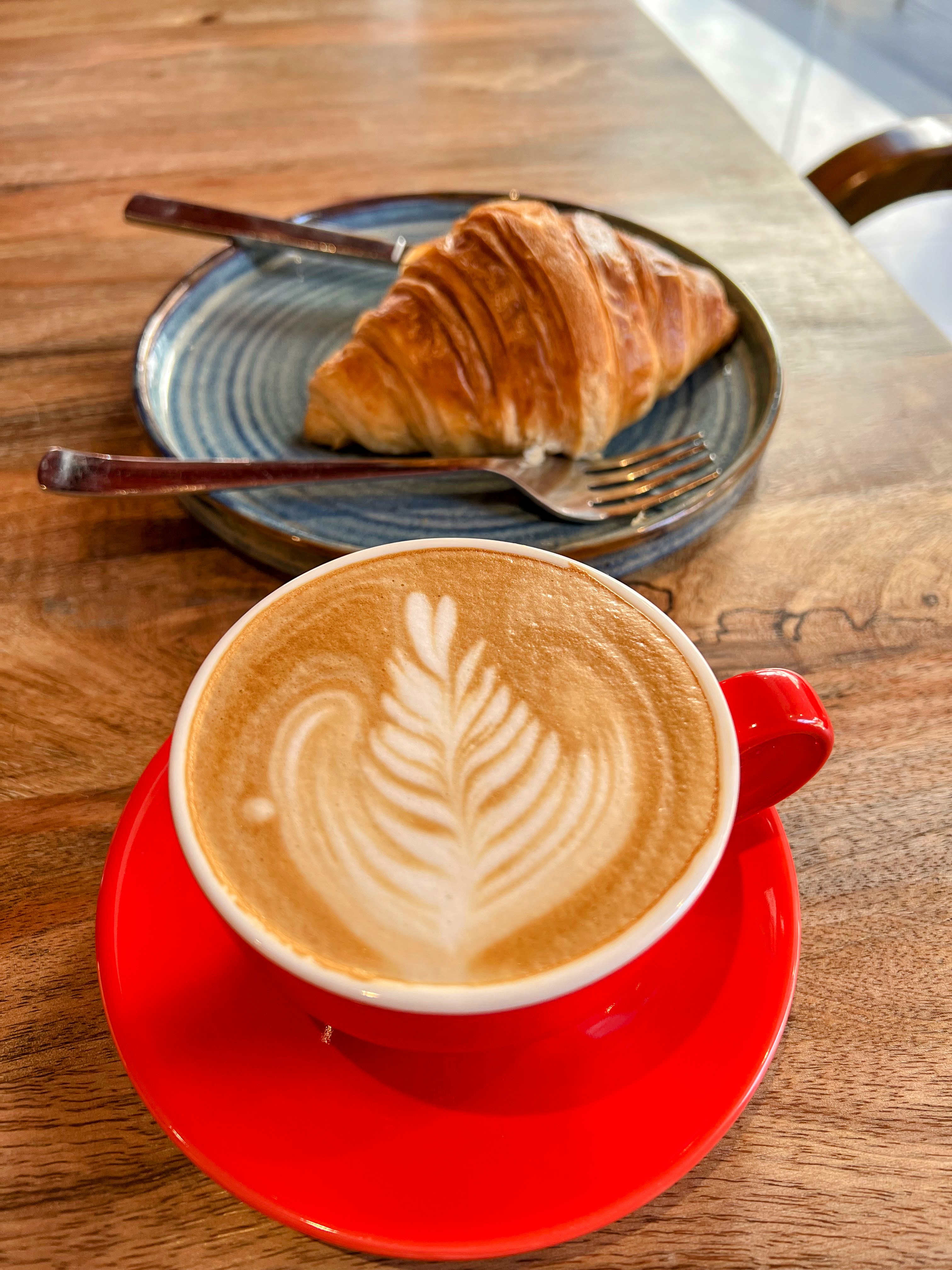 An overhead view showing a red cup of latte with a detailed fern-like latte art design, sitting next to a separate blue plate holding a golden-brown croissant and a fork.
