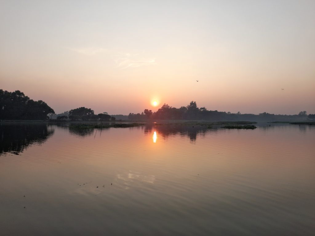 A serene sunset over a calm lake, with the orange sun reflecting on the water and trees in silhouette. Photo taken in Harbaid, Gazipur.