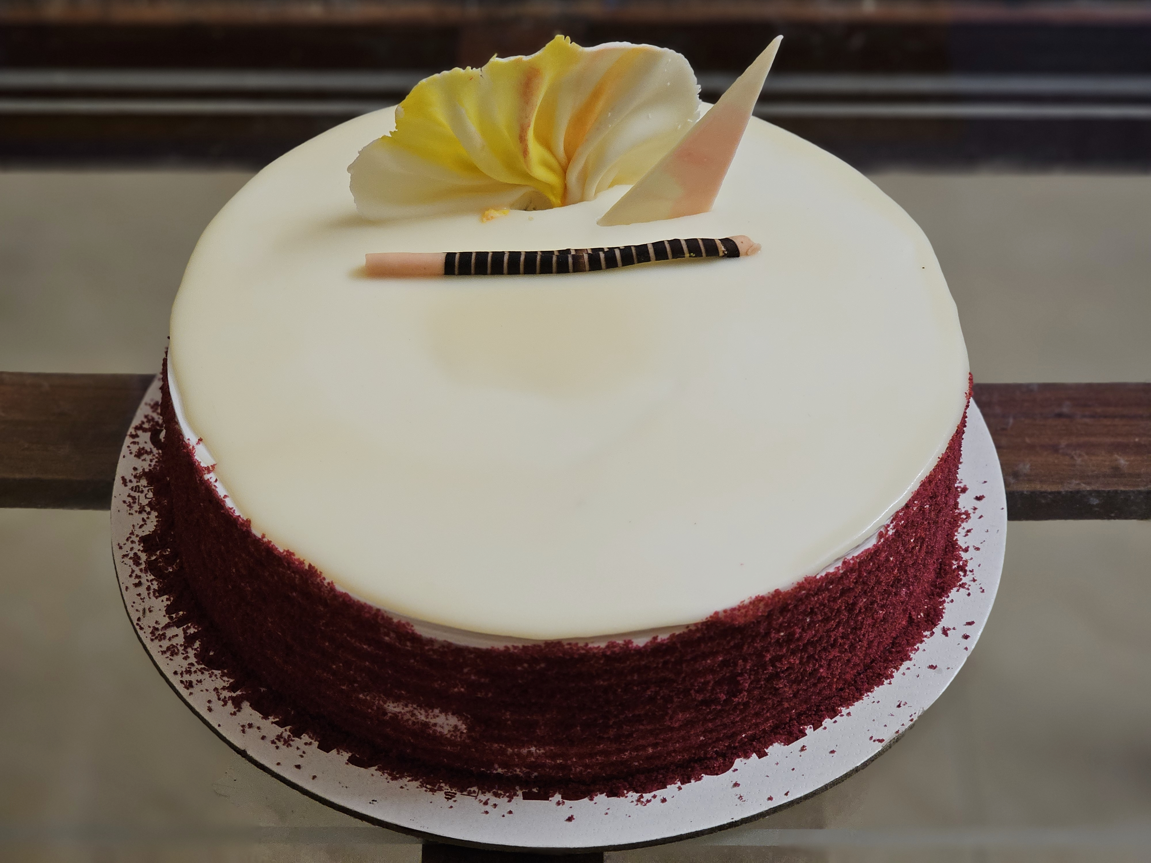 A smooth white-frosted cake with red velvet crumbs on the side, decorated with chocolate and a flower on top, was captured in Perumanna, Kozhikode, Kerala.