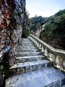 A stone staircase winds upward alongside a rocky cliff, flanked by greenery.