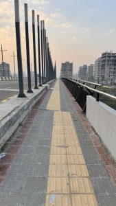 A tiled walkway with yellow tactile paving stretches along the side of a bridge with a metal railing.
