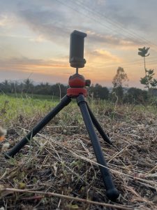 A small tripod with a red and black design is positioned on the ground, with its legs spread out. In the background, a scenic view showcases a sunset with soft clouds and a gradient of colors in the sky. 