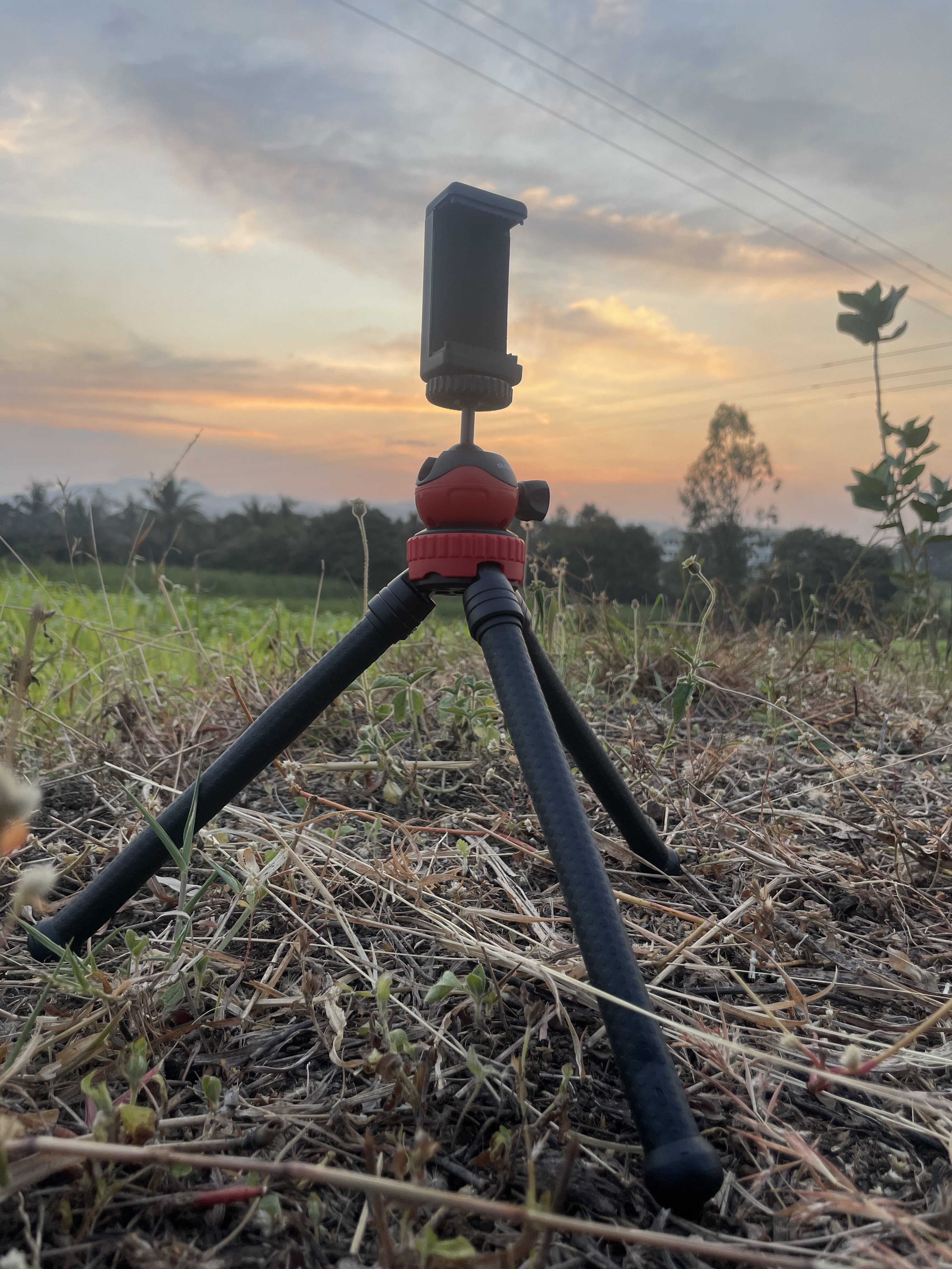 A small tripod with a red and black design is positioned on the ground, with its legs spread out. In the background, a scenic view showcases a sunset with soft clouds and a gradient of colors in the sky. 