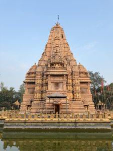 A large, intricately carved temple stands majestically, adorned with detailed sculptures and a tall spire reaching into a clear blue sky. The temple is surrounded by water, reflecting its ornate architecture. The structure features ornate motifs and carved panels, showcasing artistic craftsmanship.