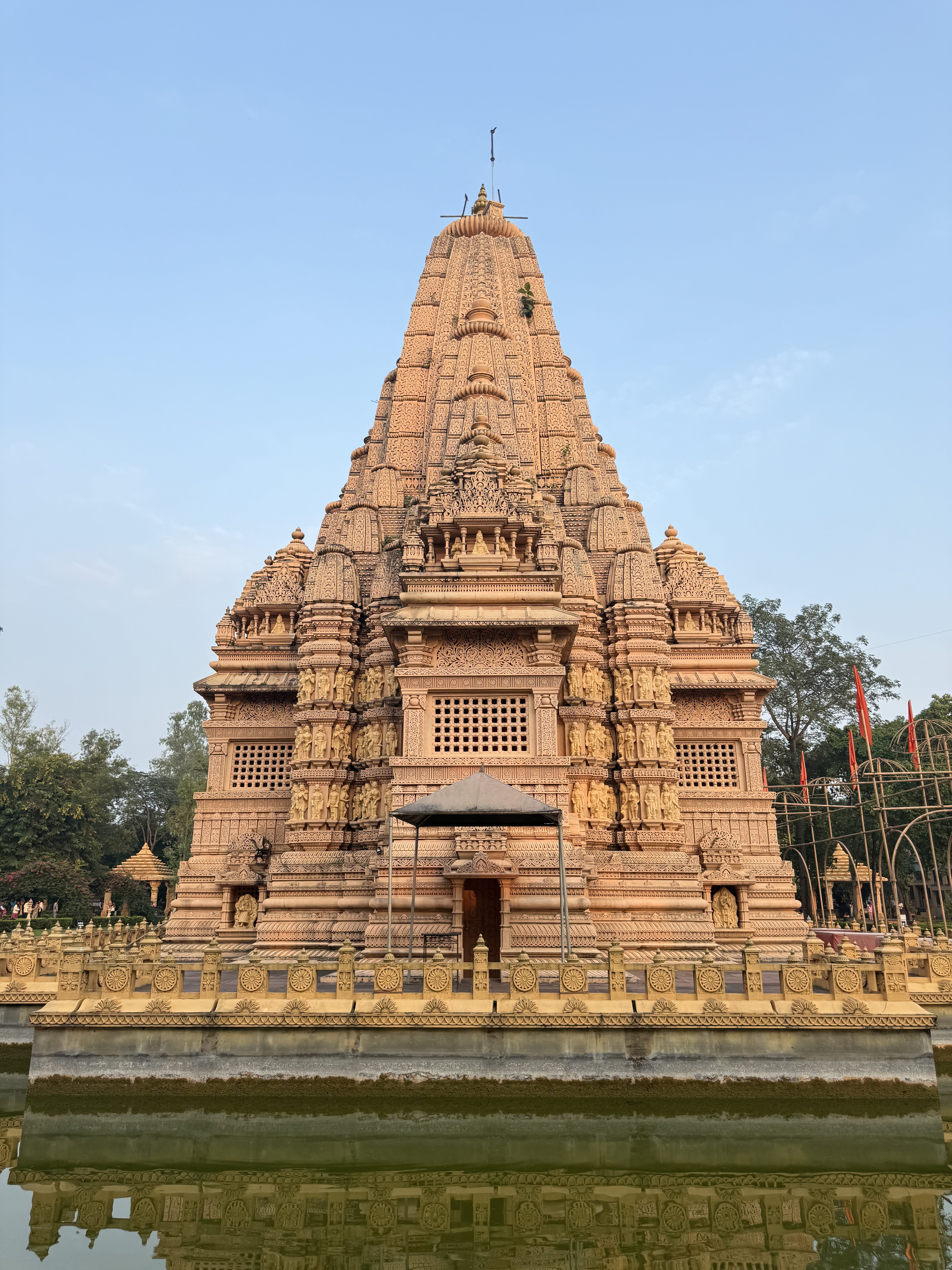 A large, intricately carved temple stands majestically, adorned with detailed sculptures and a tall spire reaching into a clear blue sky. The temple is surrounded by water, reflecting its ornate architecture. The structure features ornate motifs and carved panels, showcasing artistic craftsmanship.
