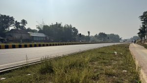 A wide paved highway stretches into the distance past small houses and green trees under a hazy bright sky.

