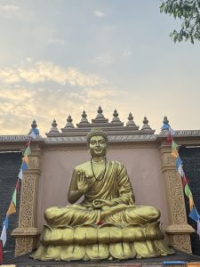 A large golden statue of a seated Buddha, positioned on a lotus pedestal, with one hand raised in a gesture of peace or blessing. The statue is located against an ornate backdrop featuring decorative stonework and a row of finial elements at the top. Colorful prayer flags hang nearby, fluttering gently in the breeze, while a soft, cloudy sky is visible in the background.