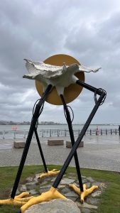 A large, artistic sculpture resembling a bird or shell, elevated on four long, slender black legs with yellow webbed feet. 
