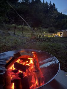 An image of a campfire. There is a metal fire pit with glowing red and partially burned wood. The background is grassy with a tent at a distance.