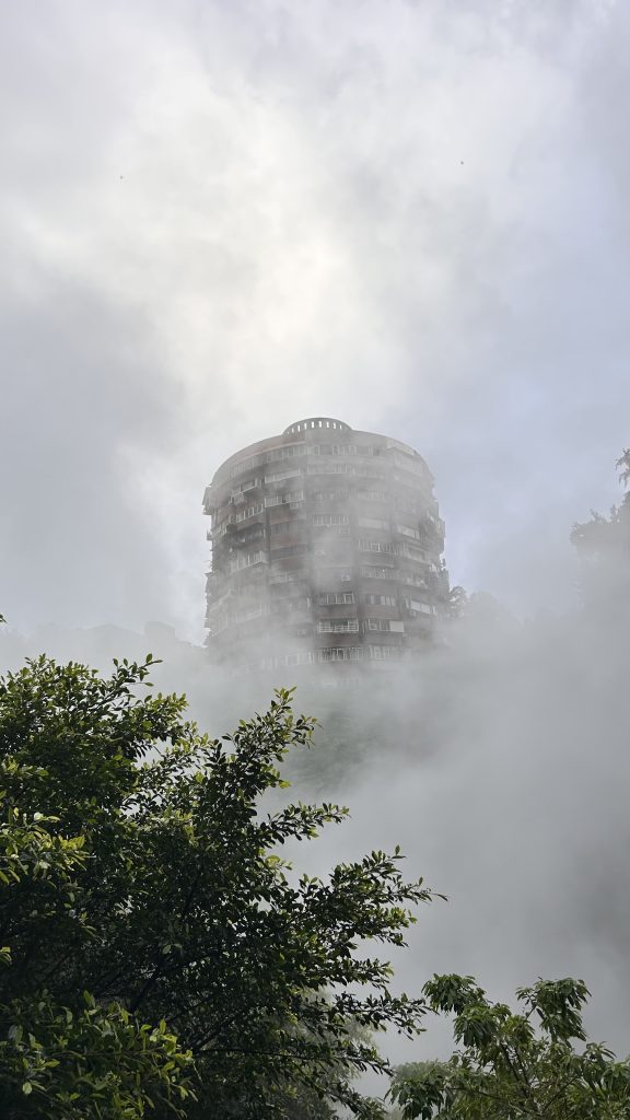 A tall, cylindrical building partially obscured by mist rises above lush greenery.