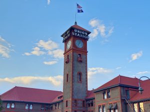 A long view of the Union Station clock tower in downtown Portland, standing tall under a bright blue sky with soft evening clouds. 