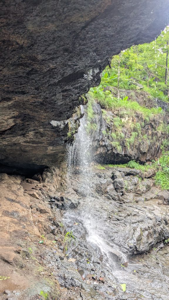 A small waterfall cascades from a rocky overhang into a rocky area below, surrounded by lush green vegetation.