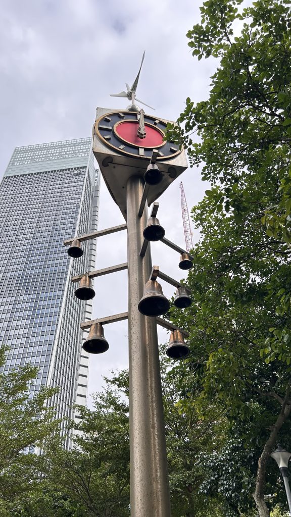 A tall, modern clock tower stands in a leafy urban environment, featuring a circular clock face with red accents, and a small wind turbine at the top.