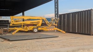 A yellow hydraulic lift is positioned on a cobblestone surface near a large, black shipping container. 