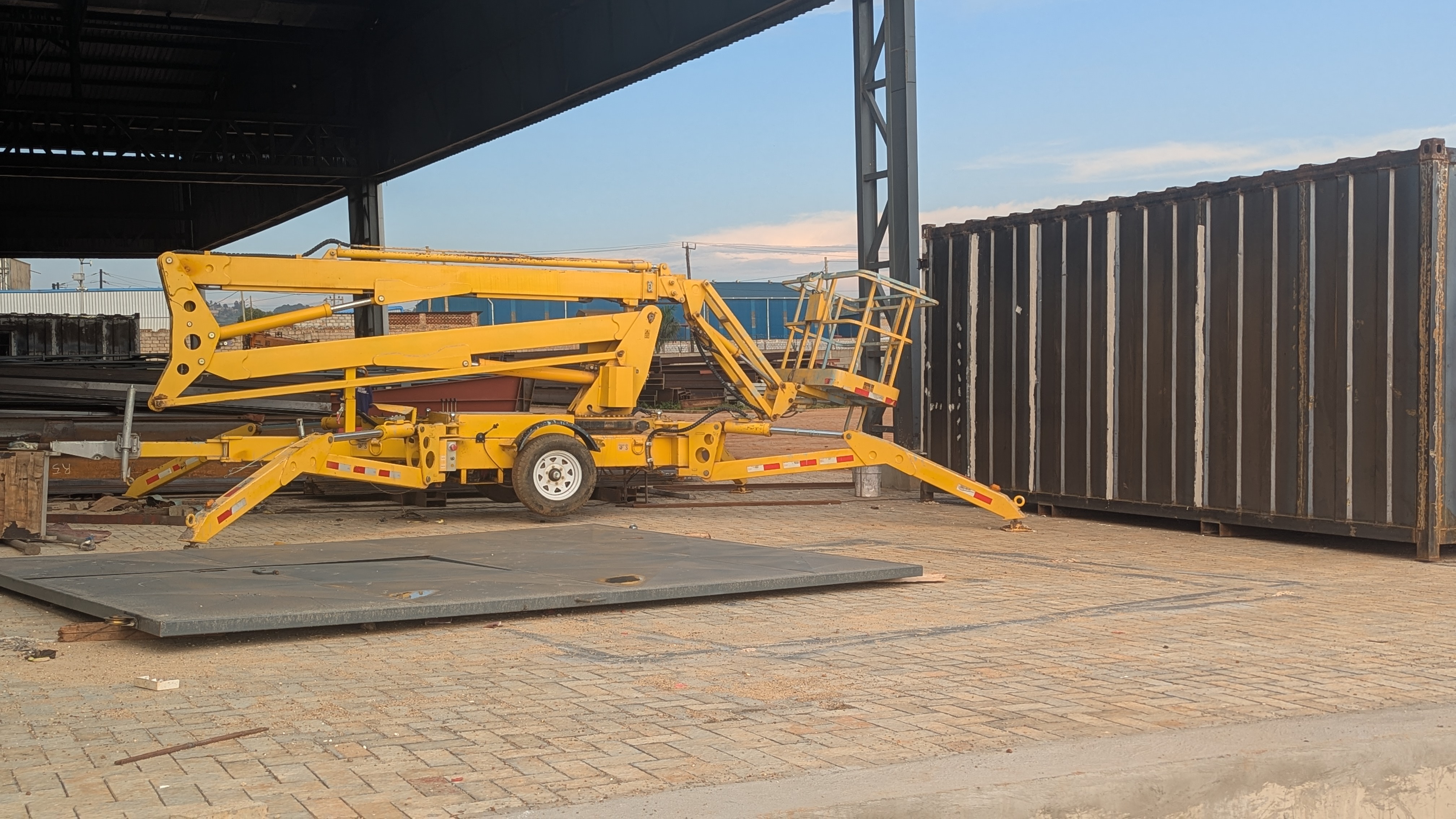 A yellow hydraulic lift is positioned on a cobblestone surface near a large, black shipping container. 